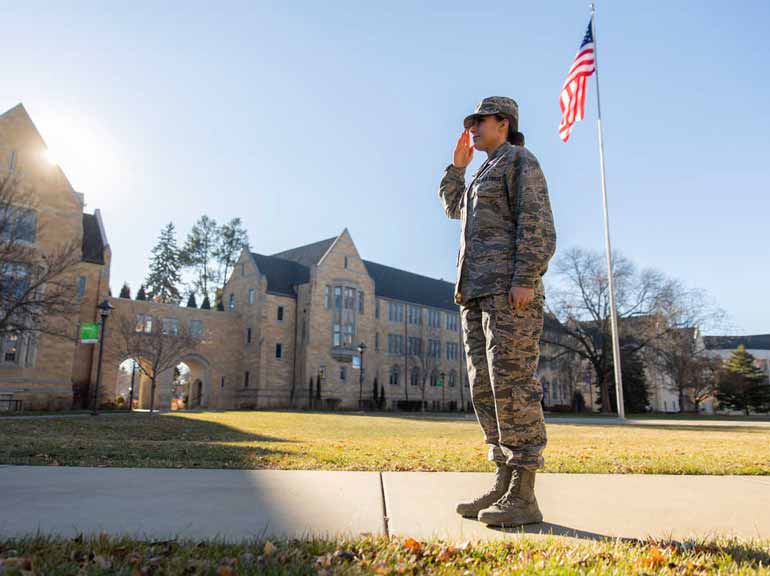 Student in military uniform salutes by flag.